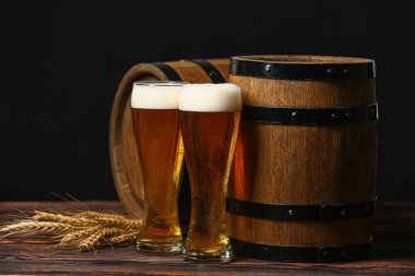 Glasses of fresh beer with spikelets and barrels on table against dark background. Oktoberfest celebration