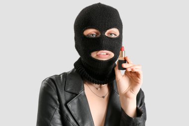 Young woman in balaclava holding red lipstick on light background