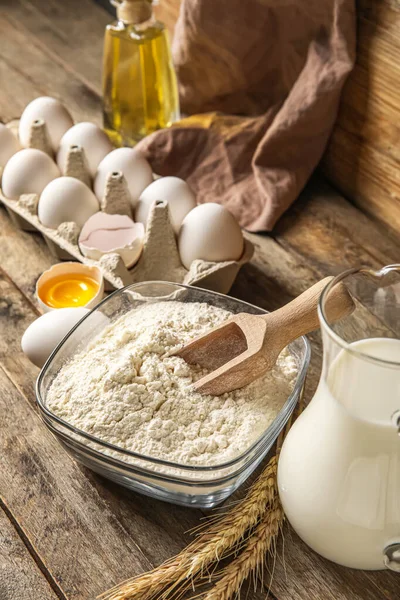 Bowl with flour, eggs and jug with milk on wooden table