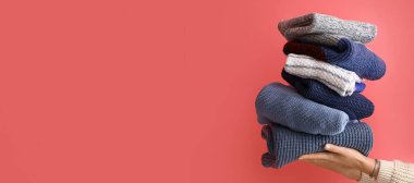 Female hands with stack of warm knitted sweaters on color background with space for text