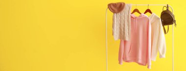 Rack with warm female clothes and accessories on yellow background with space for text