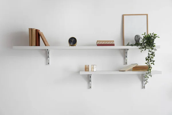 Shelves with books, frame, alarm clock and houseplant hanging on light wall