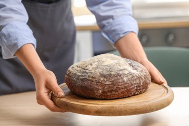 Woman putting board with fresh bread onto table in kitchen, closeup