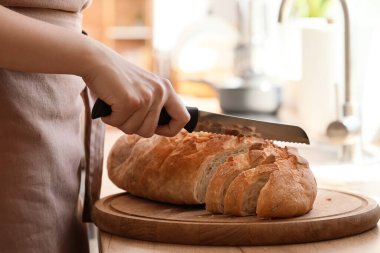 Woman cutting fresh bread on table in kitchen, closeup