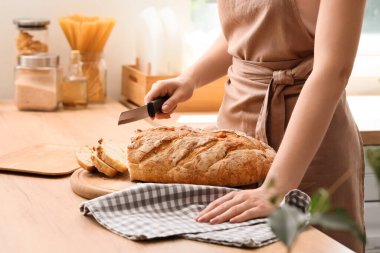 Woman cutting fresh bread on table in kitchen, closeup
