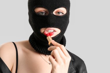 Young woman in balaclava applying red lipstick against light background