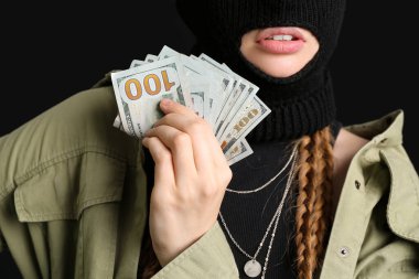 Young woman in balaclava holding money on black background, closeup