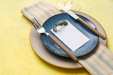Dinnerware with blank card and eucalyptus branch on yellow table, closeup