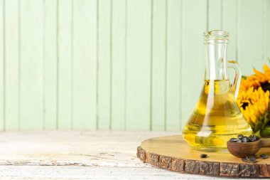 Board with jug of oil, spoon and sunflower seeds on table near green wooden wall