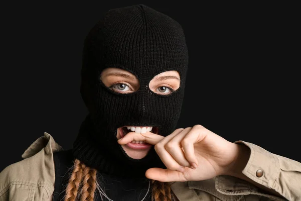 Young woman in balaclava biting finger on black background