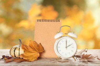 Alarm clock with notebook, autumn leaves and pumpkin on wooden table outdoors