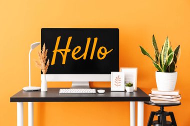Modern computer with word HELLO on screen at workplace near orange wall 