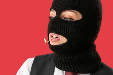 Portrait of young woman in balaclava with burning match in mouth on red background