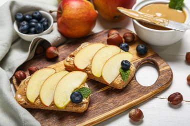 Board of tasty sandwiches with nut butter, apples and hazelnuts on light wooden background, closeup