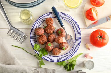 Plate with tasty meat balls, vegetables, oil and tongs on white wooden background