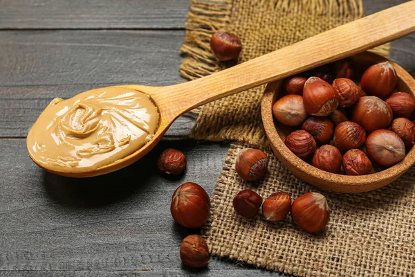 Spoon with hazelnut butter and plate of nuts on dark wooden background, closeup