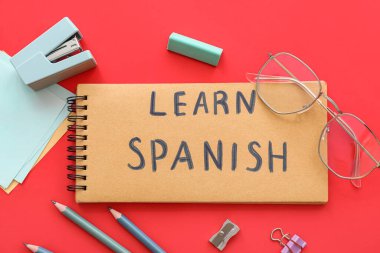 Notebook with text LEARN SPANISH, eyeglasses and stationery supplies on red background