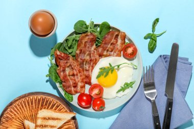 Plate with tasty fried egg, bacon, tomatoes and greens on color background