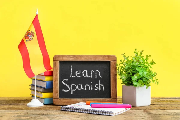 Chalkboard with text LEARN SPANISH, flag, stationery and flowerpot on table against yellow background