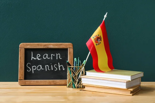 Chalkboard with text LEARN SPANISH, flag and stationery on table near green wall