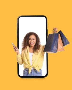 Beautiful African-American woman with bags looking out of smartphone screen on yellow background. Online shopping concept