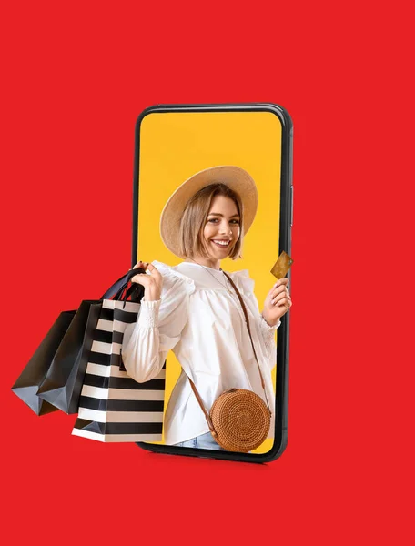 Beautiful young woman with bags and credit card looking out of smartphone screen on red background. Online shopping concept