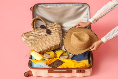 Woman putting hat into suitcase with clothes and beach accessories on pink background