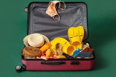 Open suitcase with beach accessories and passport on green background