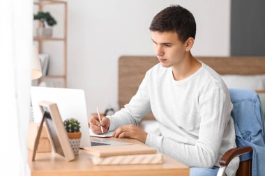 Teenage boy studying with laptop at table in bedroom