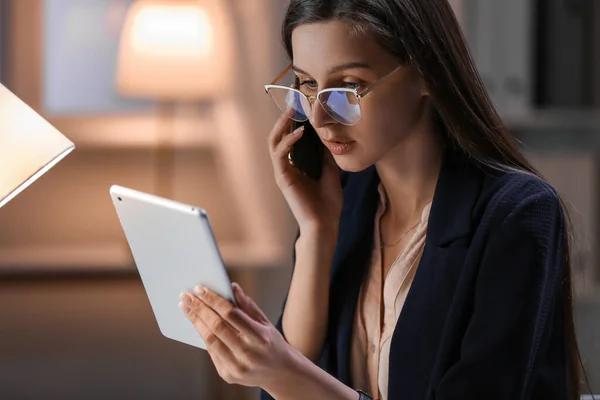 Beautiful businesswoman with tablet computer talking by mobile phone in office at night