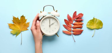 Female hand with alarm clock and autumn leaves on light blue background