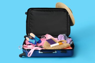 Open suitcase with clothes, beach accessories and passport on blue background
