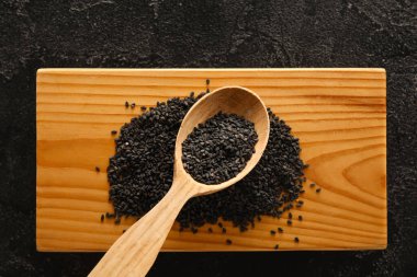 Wooden board with spoon of black sesame seeds on dark background, closeup