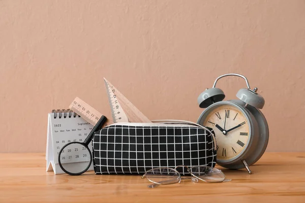 Pencil case with school stationery, eyeglasses, calendar and alarm clock on table against beige background