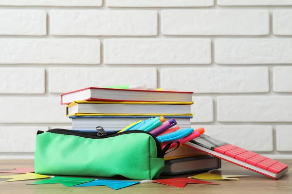 Green pencil case with school stationery and books on table near white brick wall