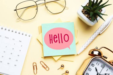 Sticky note with word HELLO, stationery, glasses, alarm clock and calendar on yellow background
