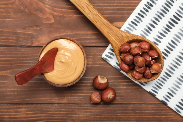 Bowl of tasty nut butter and spoon with hazelnuts on wooden background