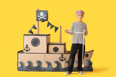 Korsan gibi giyinmiş, sarı arka planda halat ve karton gemi olan küçük bir çocuk.