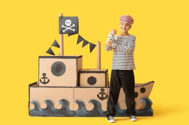 Korsan gibi giyinmiş, sarı arka planda halat ve karton gemi olan küçük bir çocuk.