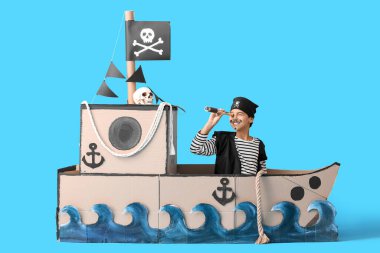 Korsan gibi giyinmiş, dürbünlü, mavi arka planda karton bir gemide.