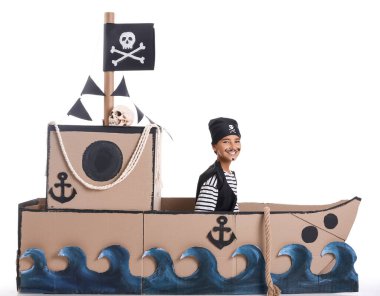 Beyaz arka planda karton bir gemide korsan gibi giyinmiş küçük bir çocuk.