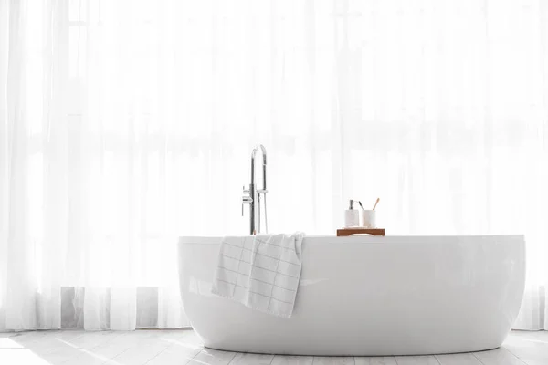 Modern bathtub with tray near window in light bathroom