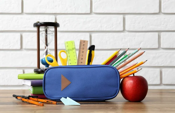 Blue pencil case with school stationery, apple and hourglass on table near white brick wall