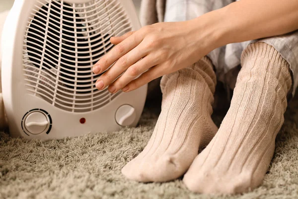 Woman warming near electric fan heater on carpet at home, closeup