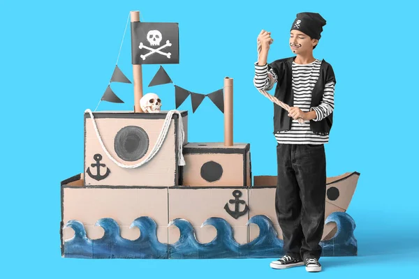 Little boy dressed as pirate with rope and cardboard ship on blue background