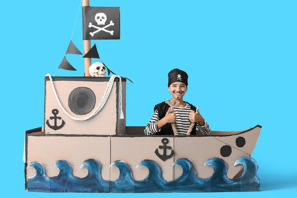 Little boy dressed as pirate with rope in cardboard ship on blue background