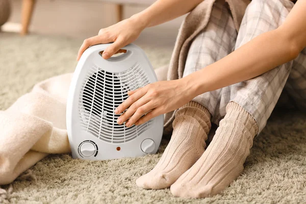 Woman warming near electric fan heater on carpet at home, closeup