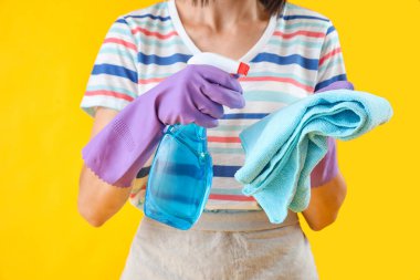 Beautiful woman with bottle of detergent and rag on yellow background