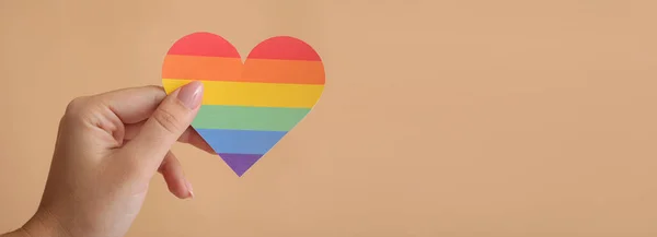 Female hand holding rainbow heart on beige background with space for text. LGBT concept