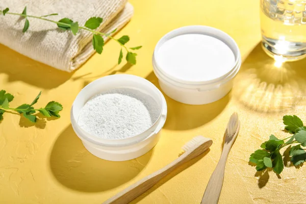 Jars of tooth powder with brushes and plant branches on yellow background